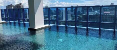 A rooftop infinity pool with clear blue water, surrounded by a glass barrier, and a city skyline in the background.