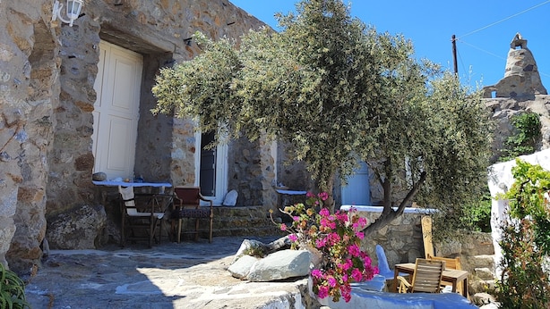 A stone building with a white door, a small table with chairs, and a tree with pink flowers. - Artemis cottage Mykonos (Mikonos)