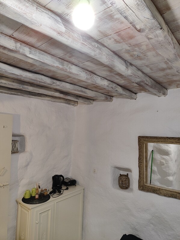 Una habitación con pared blanca, techo de madera, un armario con objetos encima, un espejo en marco y una pequeña estantería con un objeto decorativo.