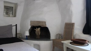 Huoneessa on sänky, pieni pöytä, jossa on kulho, rottinkituoli, takka ja seinälle kiinnitetty hylly, jonka päällä on patsas.