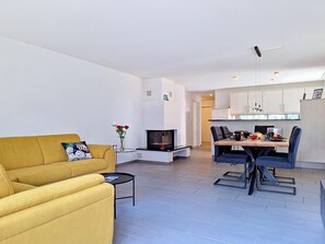 A modern living room with a yellow sofa, a small coffee table, and a fireplace. There is a dining area with a table and chairs. The kitchen is visible in the background with white cabinets. - Sans Souci O2 by Interhome (Grindelwald)