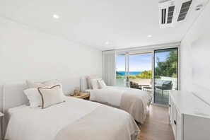 A modern bedroom with two beds, a large window with a view, and a ceiling fan. - Terrigal Apartment: Panoramic Ocean Views, Hot Tub (Terrigal)