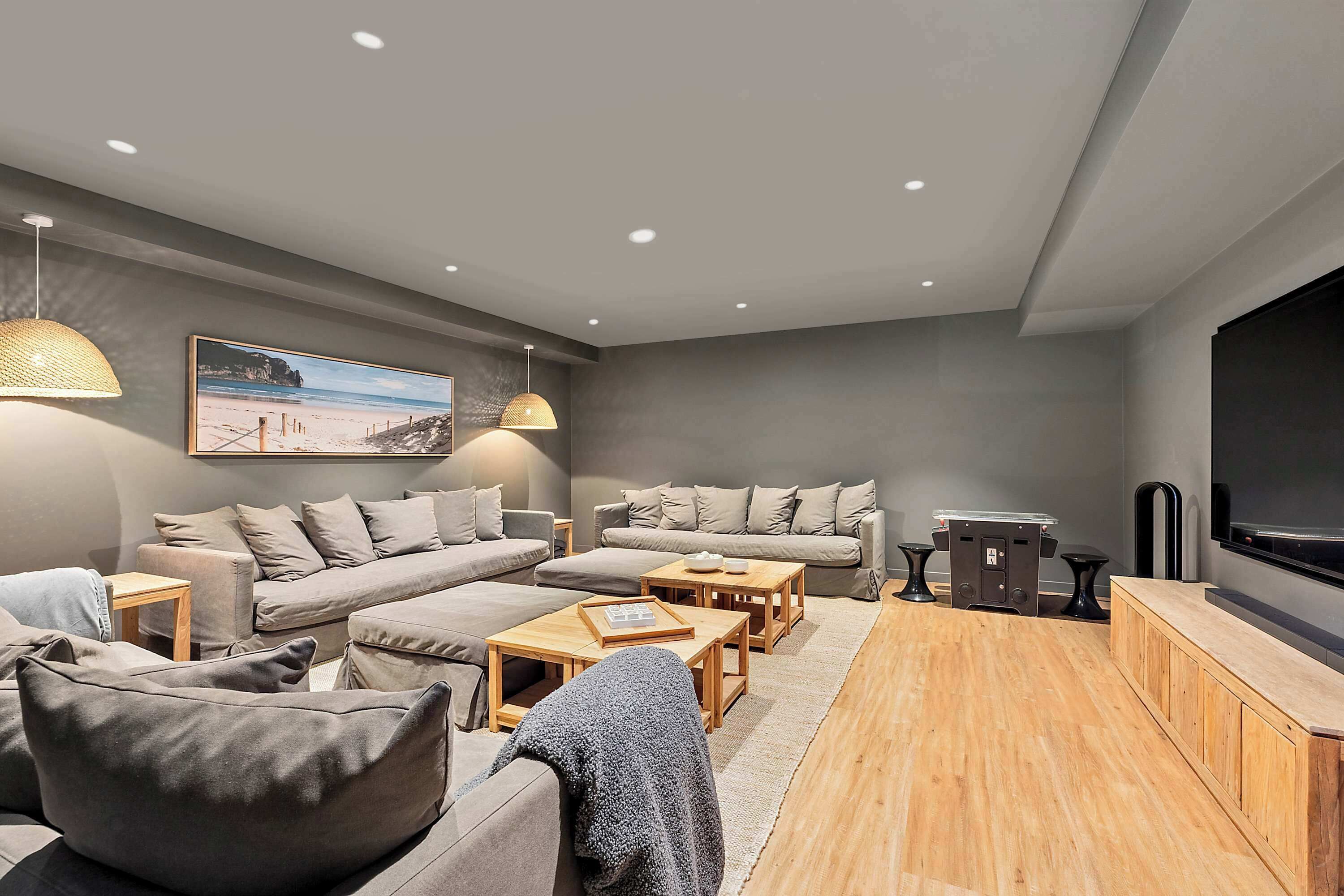 A modern living room with a sectional sofa, wooden coffee tables, a flat-screen TV, and a large painting of a beach.