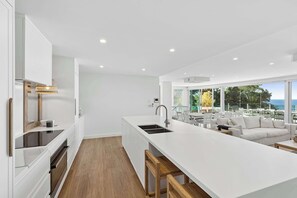 A modern kitchen with a central island, white cabinetry, and a dining area with a white sofa and table. - Terrigal Apartment: Panoramic Ocean Views, Hot Tub (Terrigal)
