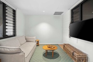 A living room with a sofa, a round wooden coffee table, a TV mounted on the wall, and a window with blinds. - Drift by Coast Hosting (Avoca Beach)