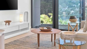 A modern living room with a TV, a round wooden table, a glass coffee table, and a view of greenery through large windows. - Avoca Lake View Luxury Apartment (Avoca Beach)