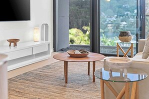 A modern living room with a TV, a round wooden table, a glass coffee table, and a view of greenery through large windows. - Avoca Lake View Luxury Apartment (Avoca Beach)