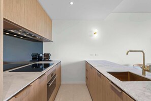 A modern kitchen with wooden cabinets, a black induction cooktop, a stainless steel range hood, a sink with a gold faucet, and a wall-mounted light. - Drift by Coast Hosting (Avoca Beach)