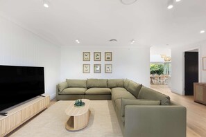 A modern living room with a large flat-screen TV, a sectional sofa, a coffee table, and framed artwork on the wall. - The Oasis by Coast Hosting (Umina Beach)