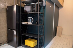 A compact kitchenette with a refrigerator, microwave, kettle, and a yellow storage bin.