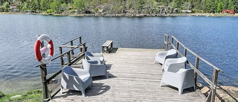 A wooden dock with white chairs and a lifebuoy, overlooking a calm lake with a forested shoreline.