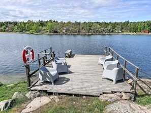 Un muelle de madera con sillas blancas y un flotador salvavidas, con vistas a un lago tranquilo y una orilla boscosa.