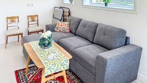 Ein modernes Wohnzimmer mit einem grauen Sofa, zwei gestreiften Sesseln, einem Holztisch mit floralem Tischläufer und einem Fenster mit Blick ins Grüne.