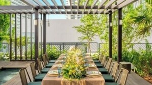 Een lange eettafel gedekt voor een maaltijd, met stoelen op een houten terras, omringd door groen.