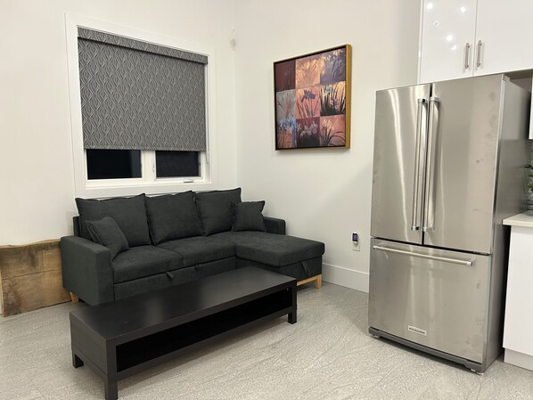 A modern kitchen with a stainless steel refrigerator, a dark grey sectional sofa, and a patterned window blind.