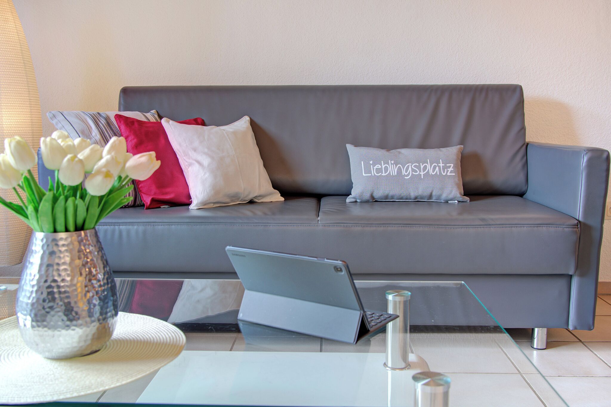 Ein graues Sofa mit einem Laptop auf einem Glastisch, eine Vase mit weißen Blumen und ein Kissen mit dem Aufdruck 'Lieblingsplatz'.