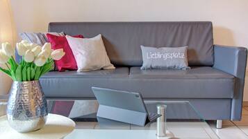 Ein graues Sofa mit einem Laptop auf einem Glastisch, eine Vase mit weißen Blumen und ein Kissen mit dem Aufdruck 'Lieblingsplatz'.
