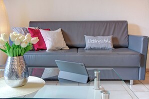 A grey sofa with a laptop on a glass table, a vase with white flowers, and a pillow with 'Lieblingsplatz' text. - Depedance house Gisela with 3 bedrooms (Bann)