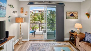 A living room with a TV, a lamp, a sofa, a coffee table, and a balcony with a view of palm trees and buildings. - Moorings Pool Overview Resort (Lantana)