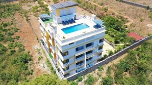 Un hotel de varios pisos con piscina en la azotea, rodeado de vegetación y un muro de piedra.