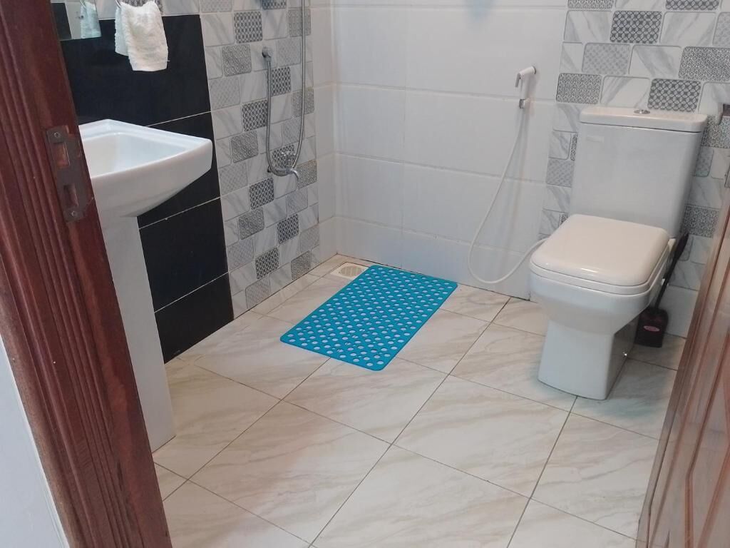 Casa de banho com lavatório, sanita e chuveiro, e um tapete azul no chão.
