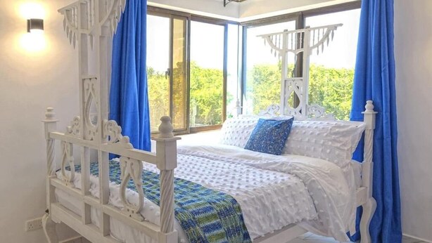 Una cama con dosel blanco con una manta estampada, cortinas azules y vistas a la vegetación a través de amplias ventanas.