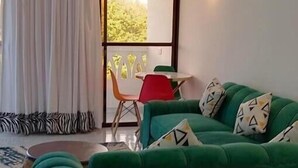 Un sofá verde con cojines estampados, una mesita redonda pequeña, una silla roja y vistas a la vegetación a través de una ventana.