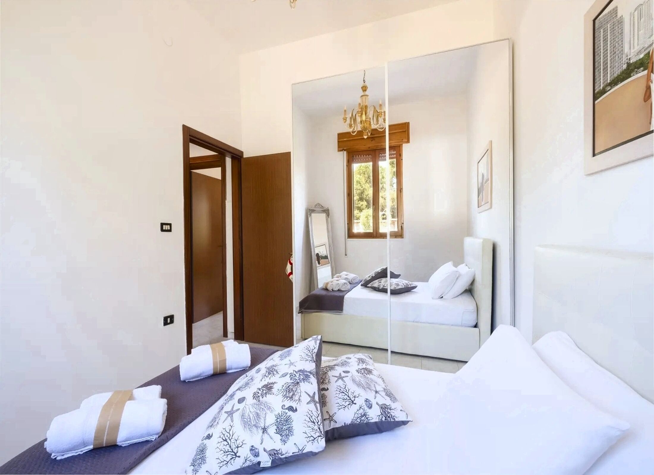 Habitación de hotel con cama, un espejo grande, una araña y una ventana con luz natural.