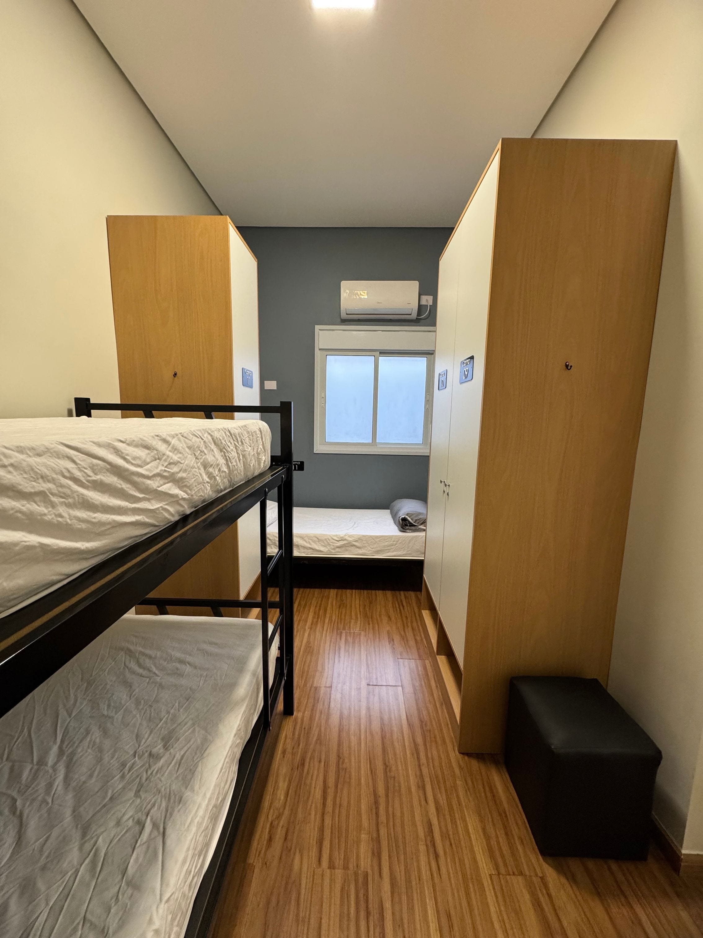 Quarto em dormitório com beliches, janela, ar-condicionado e armários de madeira.