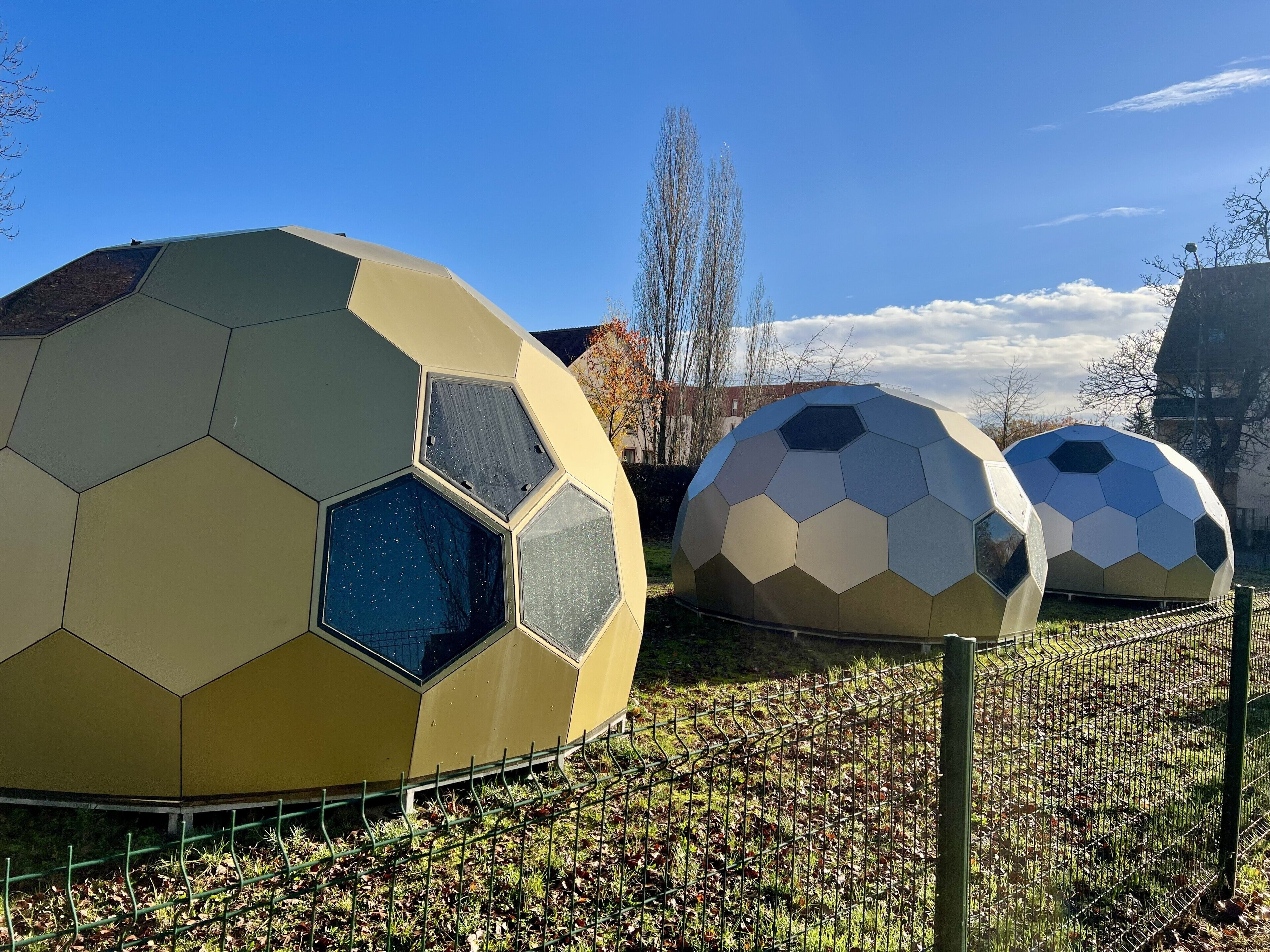 Deux structures sphériques ornées de motifs hexagonaux, entourées d’une clôture et d’arbres.
