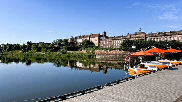 Ein ruhiger Fluss mit am Holzsteg vertäuten Booten, ein großes historisches Gebäude im Hintergrund und ein klarer, blauer Himmel.
