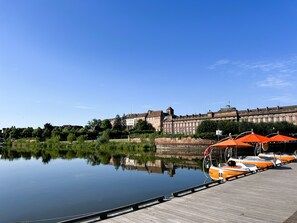 Une rivière paisible où des bateaux sont amarrés à un quai en bois, un grand bâtiment historique en arrière-plan et un ciel bleu dégagé.