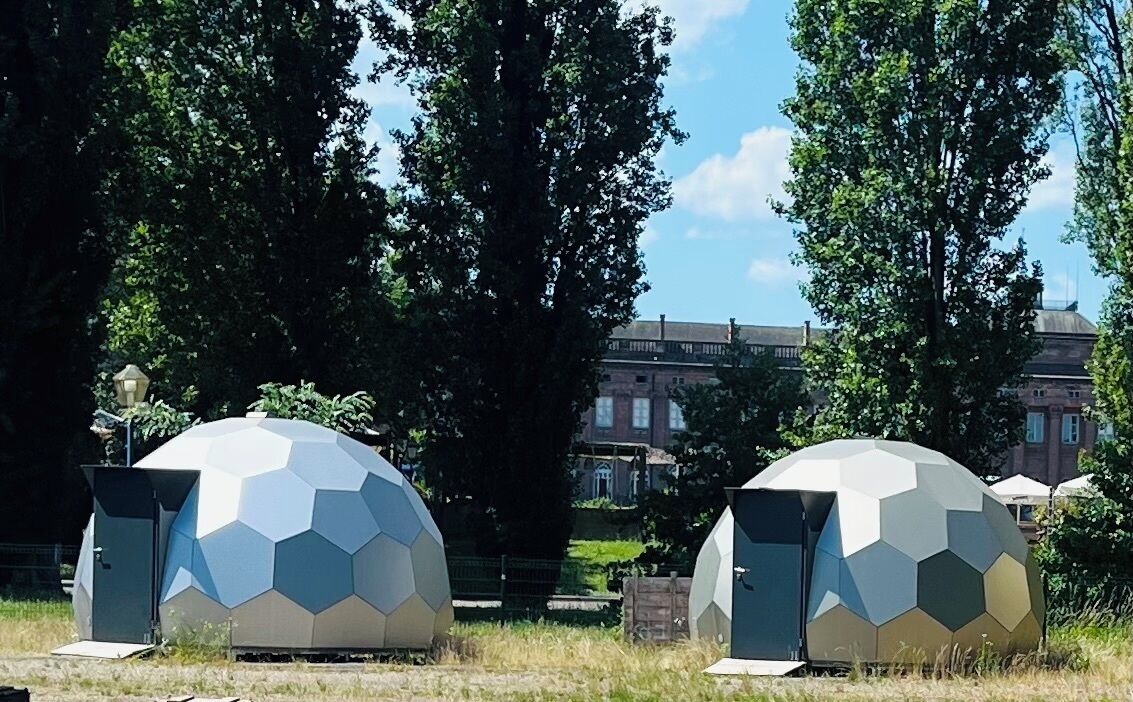 Two spherical structures with hexagonal patterns, possibly for accommodation or events, in a park with trees and a building in the background.