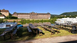 Eine Uferlandschaft mit einem großen, historischen Gebäude, einem Boot, das am Fluss liegt, und Picknicktischen, die am grünen Ufer angeordnet sind.