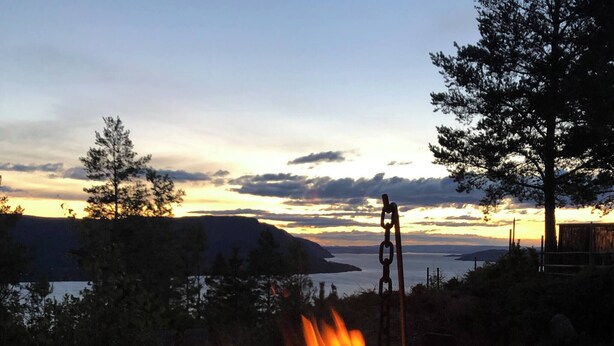 Ein Lagerfeuer mit brennenden Holzscheiten mit Blick auf ein Gewässer und Hügel.