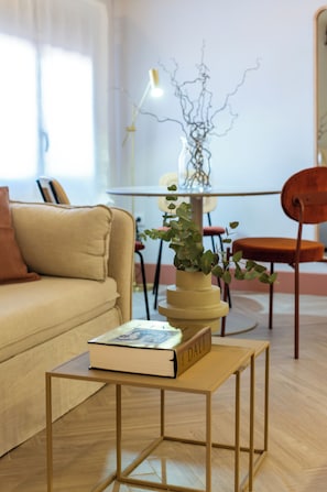 Een woonkamer met een bank, een bijzettafeltje met een boek, een plant in pot, een eettafel met stoelen en een lamp.