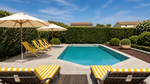 Piscina con sillas de descanso y sombrillas, rodeada de setos y plantas en macetas.