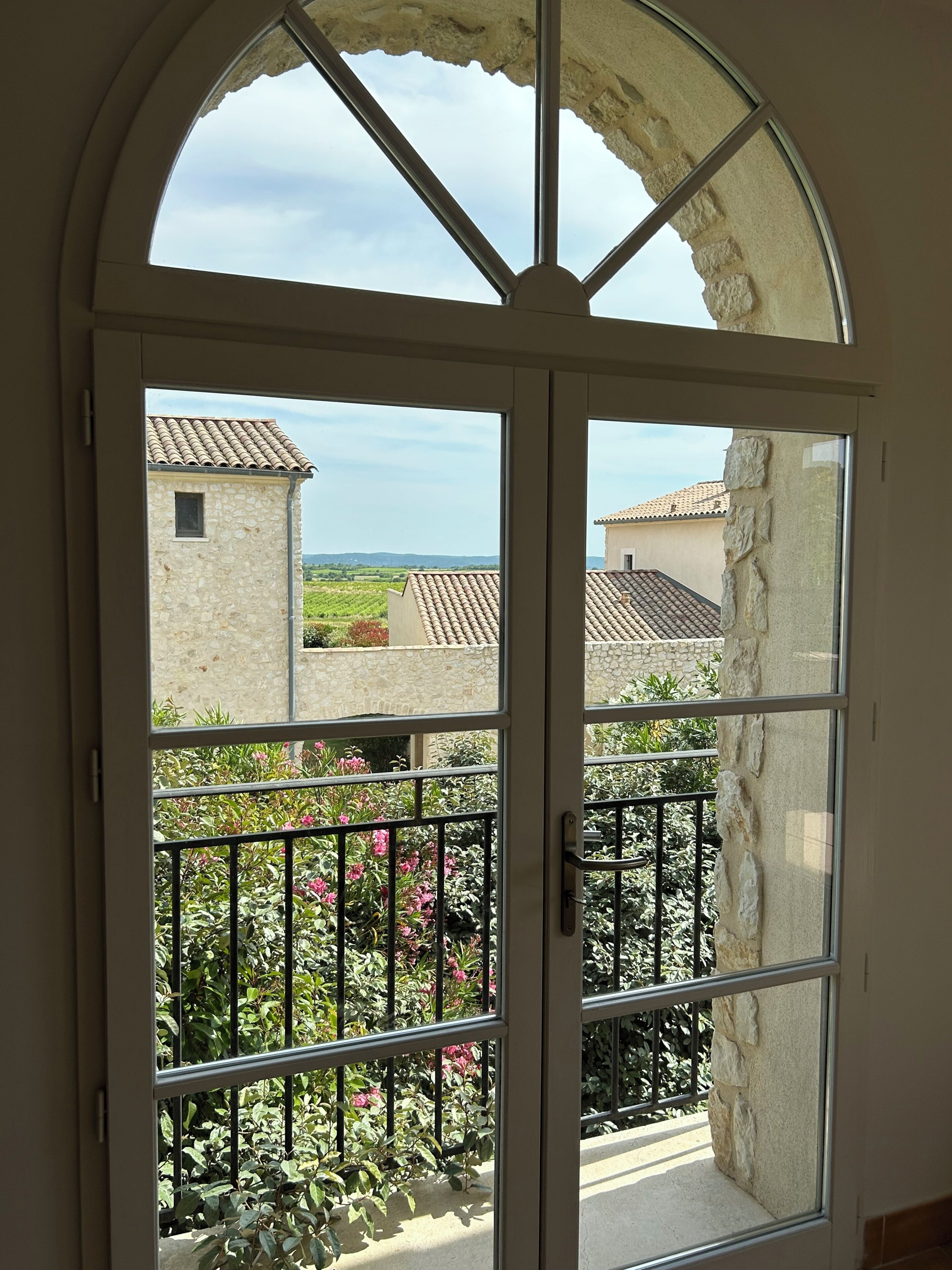 A view through a glass door with a stone wall, a balcony with metal railings, and buildings with tiled roofs in the background.