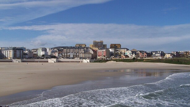 Paesaggio costiero con spiaggia sabbiosa, onde dell'oceano e una fila di edifici a più piani.