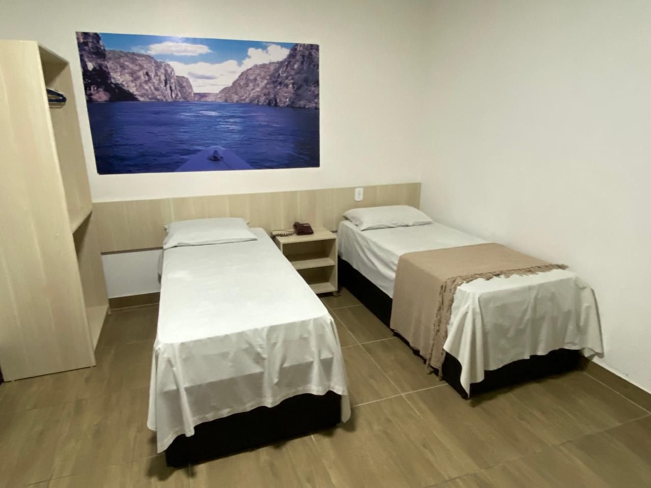 A small room with two single beds, a nightstand, and a large wall-mounted picture of a coastal landscape.