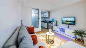 En moderne stue med sofa, tv, lille bord og et køkken i baggrunden.