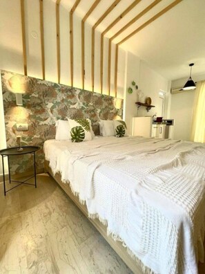 A bedroom with a stone wall, wooden beams, a bed with white bedding, a small table, and a lamp. - Eora Studios (Zakynthos)