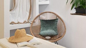 Une pièce bien rangée, avec un lit, une chaise en osier dotée d’un coussin, une corbeille suspendue et une étagère sur laquelle est posée une plante.