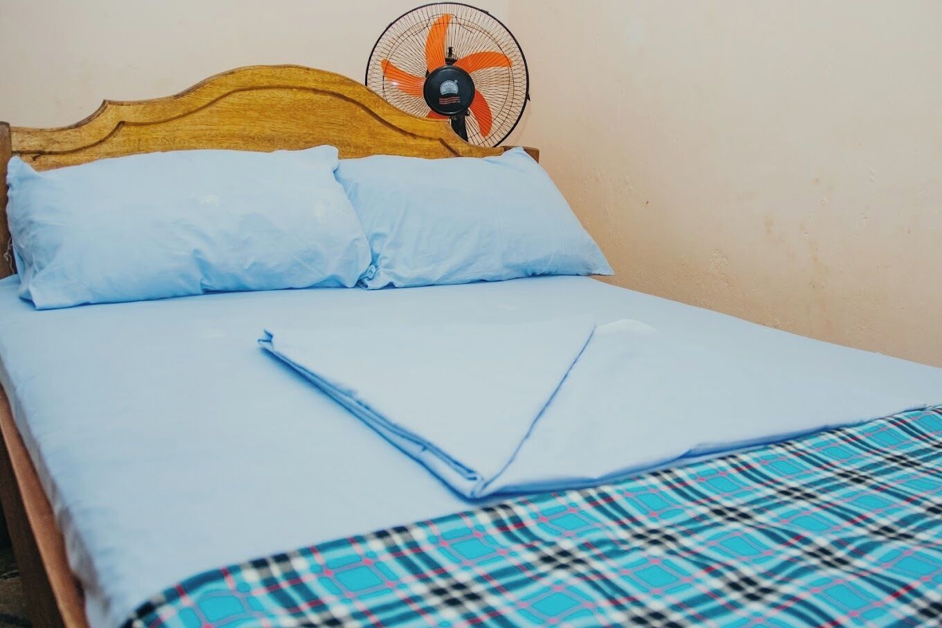 Un lit avec une tête de lit en bois, deux oreillers, une couverture pliée et un ventilateur fixé au mur.