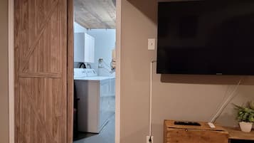 A room with a mounted TV, wooden furniture, and a kitchen area visible through an open door.