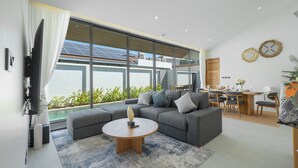A modern living room with a grey sofa, round coffee table, and large windows overlooking a pool. - Seaside New 3BR Pool Villa 2 Min Walk to Bangtao Beach, Villa Belplaya Phuket (Tambon Choeng Thale)