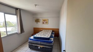 Ett litet rum med en enkel säng, ett fönster, en takfläkt och en väggmonterad luftkonditioneringsanläggning.