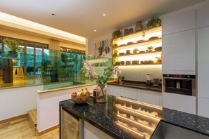 A modern kitchen with a glass dining area, a variety of plants, and a central island with a fruit bowl.