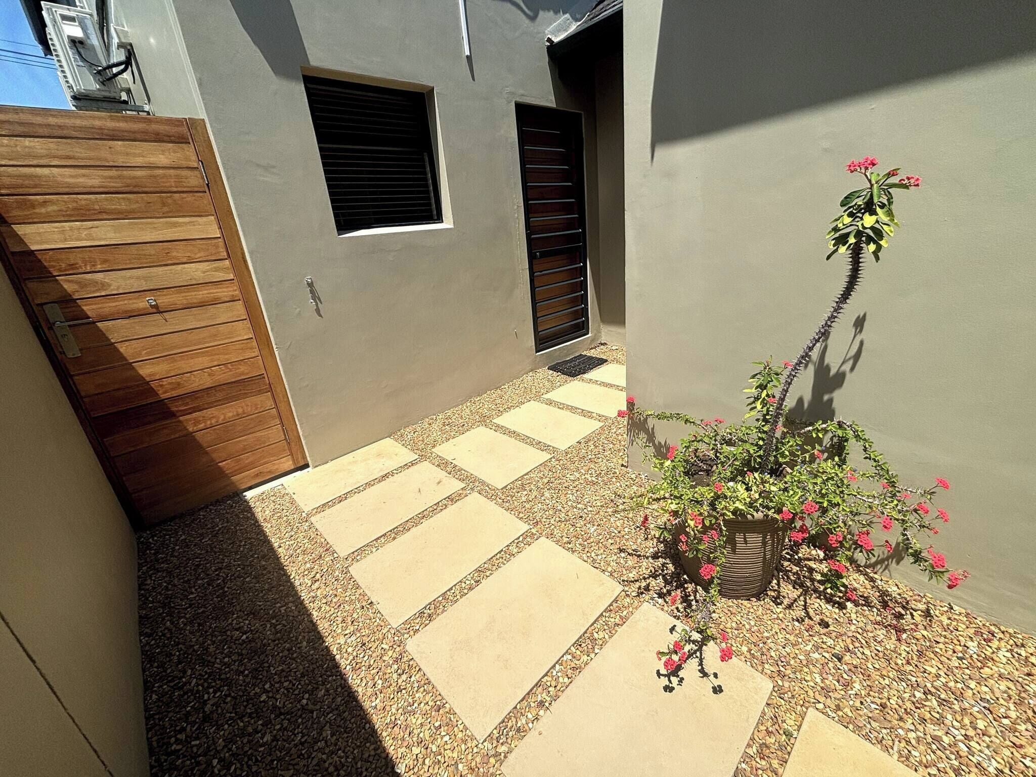 Une cour avec une porte en bois, un pot de fleurs orné de fleurs roses et un chemin pavé.