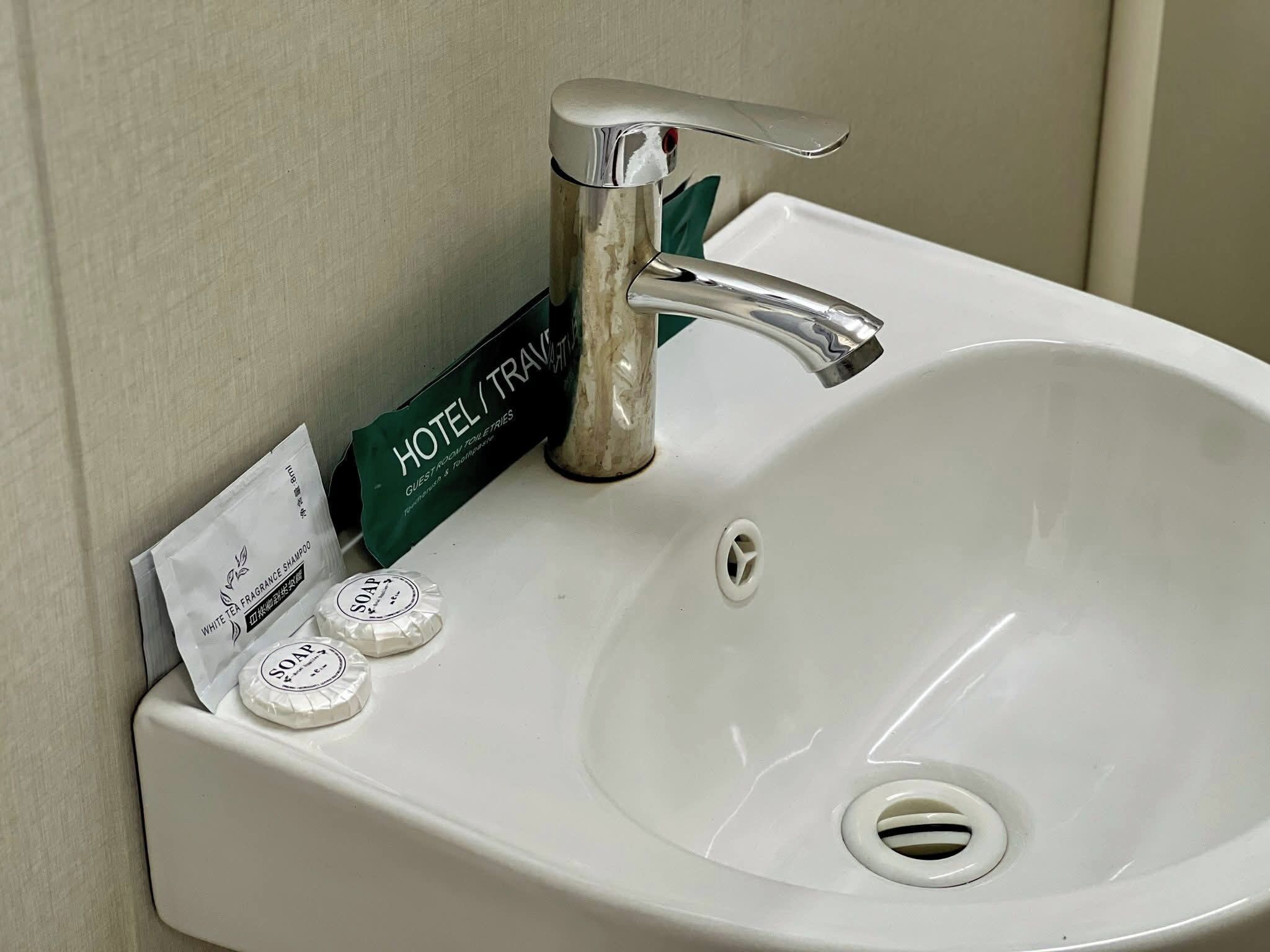 Un lavamanos blanco con grifo cromado, un cartel de hotel/viajes verde, y dos sobres de jabón.